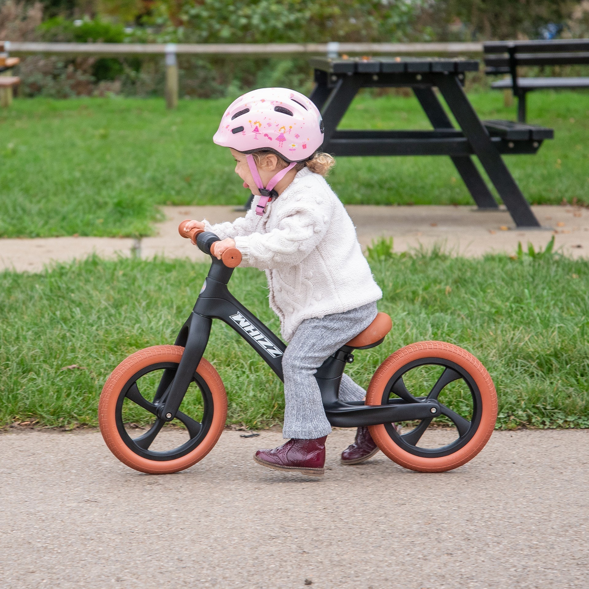 Child riding a balance bike with pink helmet in a park setting