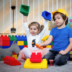 Two children playing with colorful toy building blocks and a small toy hammer in a bright indoor play area. The child on the left holds a green block above her head, and the child on the right wears a yellow hard hat while holding an orange-handled hammer.