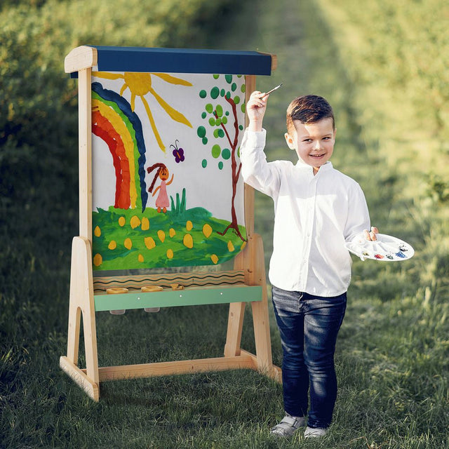 Child standing next to a colorful drawing on an easel outdoors