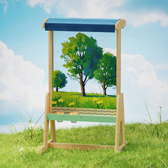 A wooden easel-style art stand with a blue top panel and a green landscape painting displayed on the front, set against a grassy field and blue sky background.