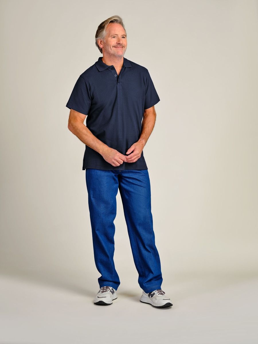 A man wearing a dark navy blue short-sleeve polo shirt and blue jeans, standing against a plain light beige studio background.