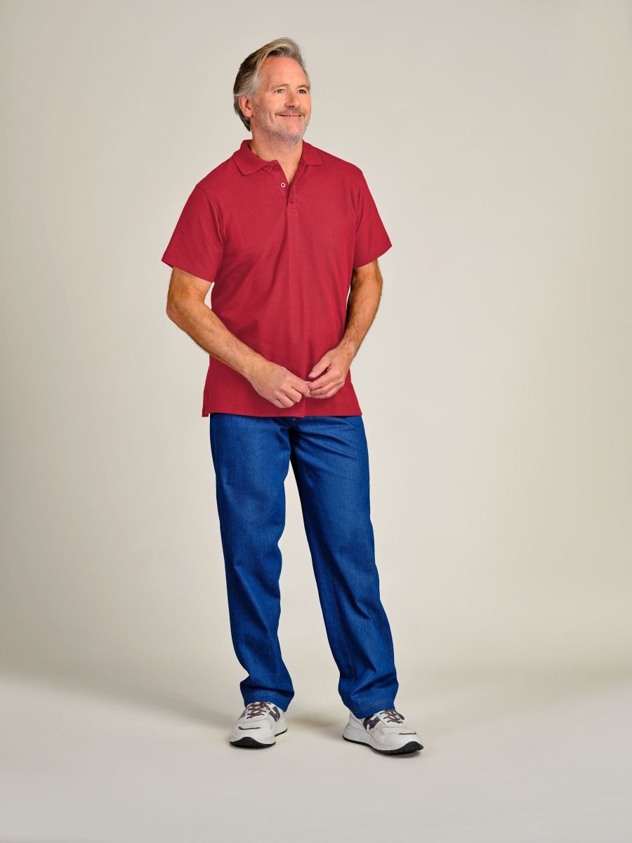 A man wearing a red short-sleeve polo shirt and blue pants, standing against a plain light beige studio background.