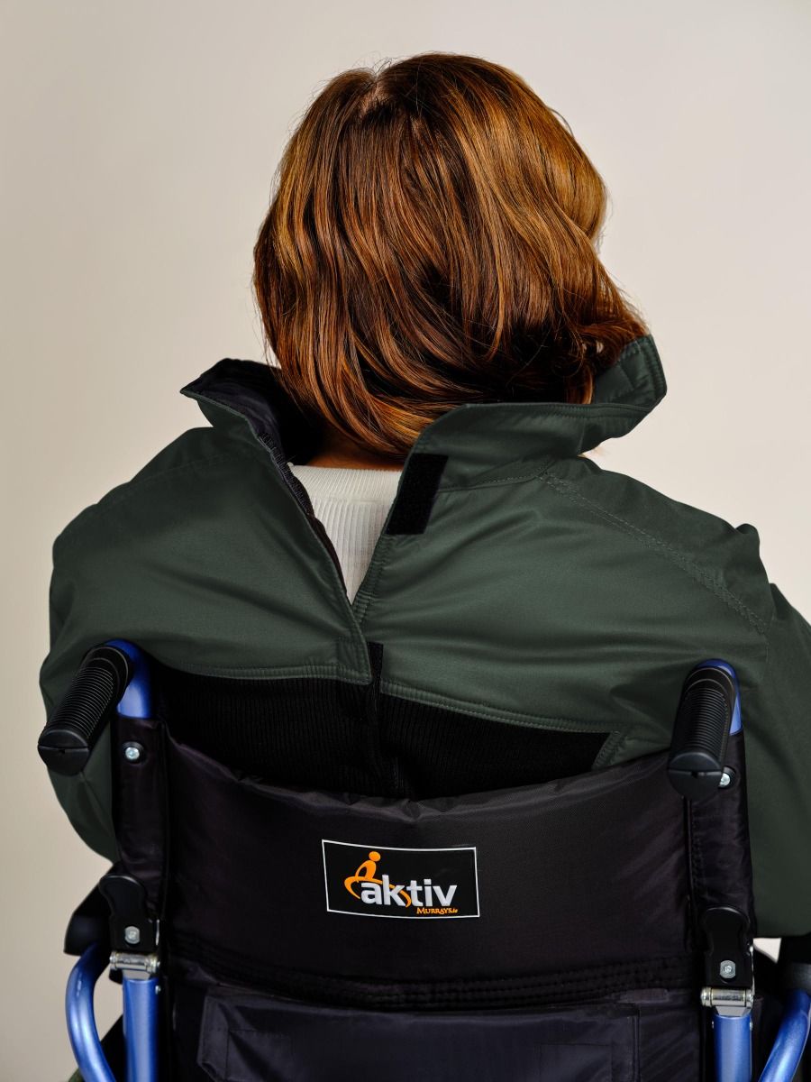 A person viewed from behind wearing a dark olive green jacket and sitting in a black Aktiv wheelchair against a plain light background.