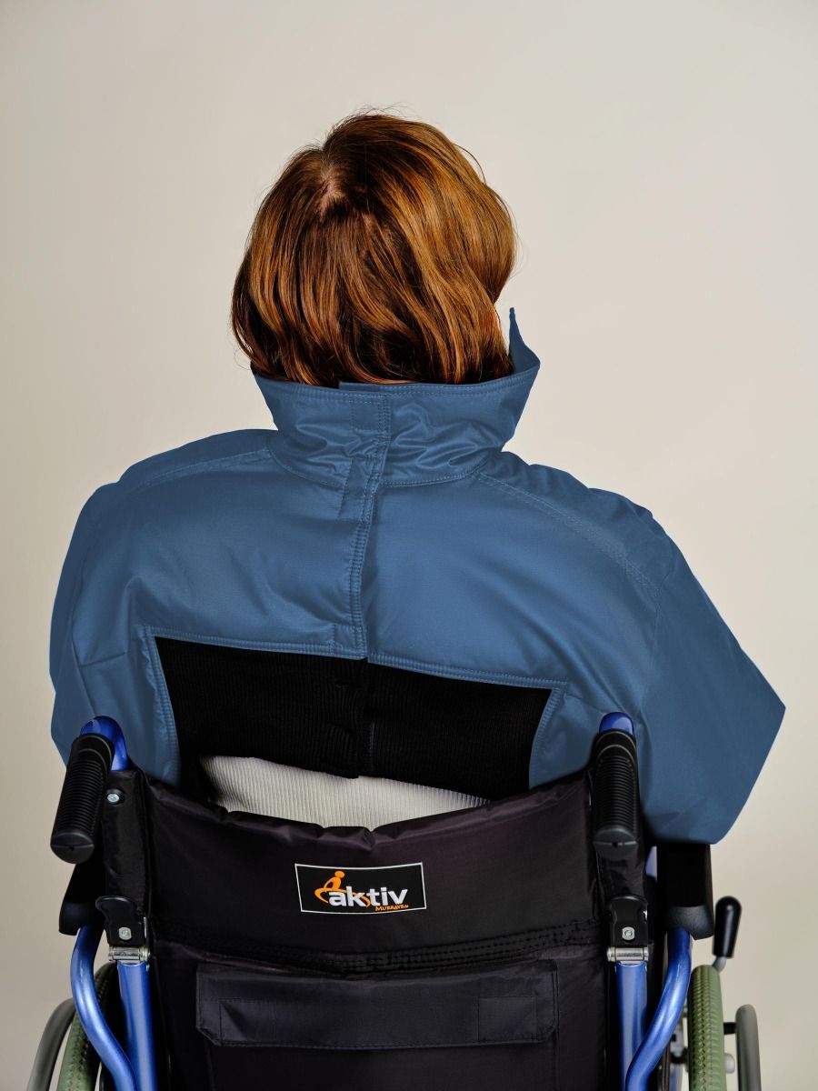 A person with short auburn hair sits in a blue and black Aktiv wheelchair, viewed from behind, wearing a blue high-collar jacket against a plain light gray background.