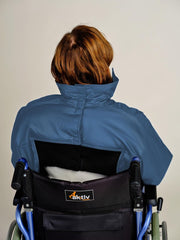 A person with short auburn hair sits in a blue and black Aktiv wheelchair, viewed from behind, wearing a blue high-collar jacket against a plain light gray background.