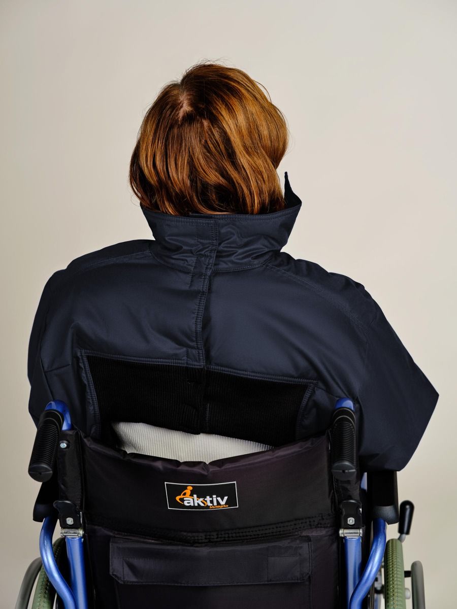 A person with short auburn hair sits in a wheelchair viewed from behind, wearing a dark navy blue high-collar jacket. The wheelchair has a black backrest with a visible 'aktiv' logo patch.