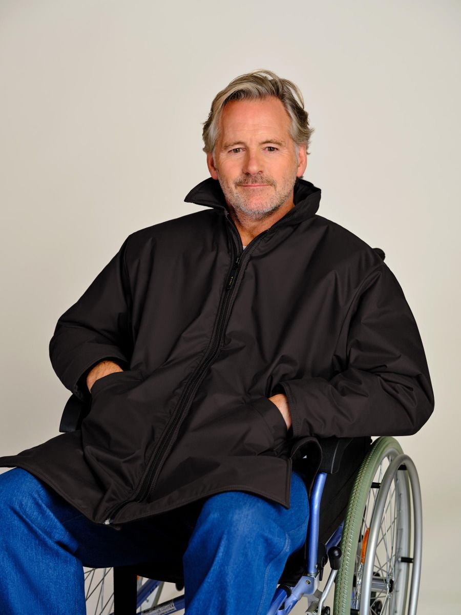 A man with gray hair and a beard sitting in a wheelchair, wearing a black zip-up jacket with a high collar and blue jeans, photographed against a plain light gray background.