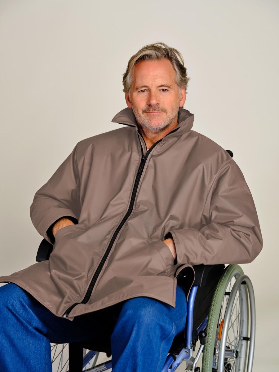 A man with gray hair and a beard sitting in a wheelchair, wearing a brown waterproof jacket with a high collar and blue jeans, against a plain light gray background.