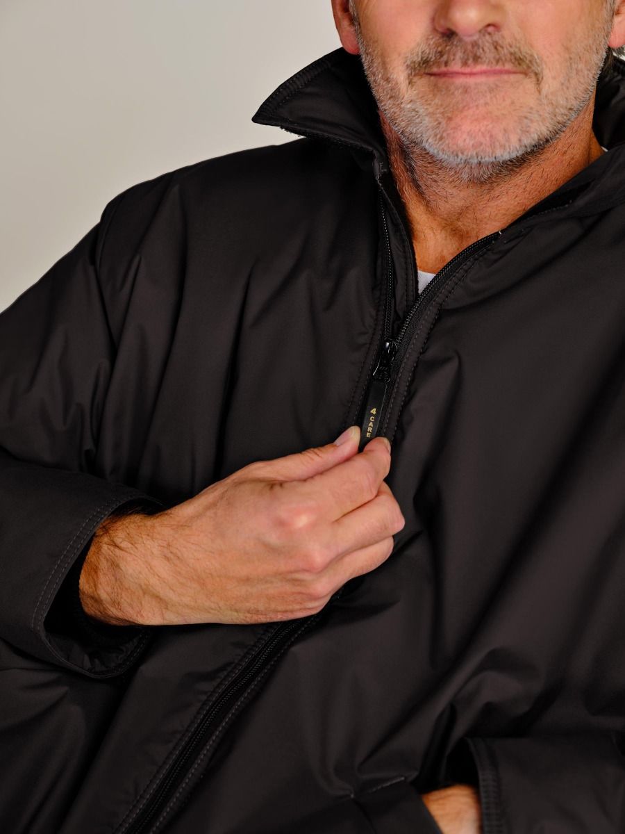 Close-up of a man wearing a black zip-up jacket, pulling the zipper with his hand. The photo is cropped from the lower face to the torso against a plain light background.