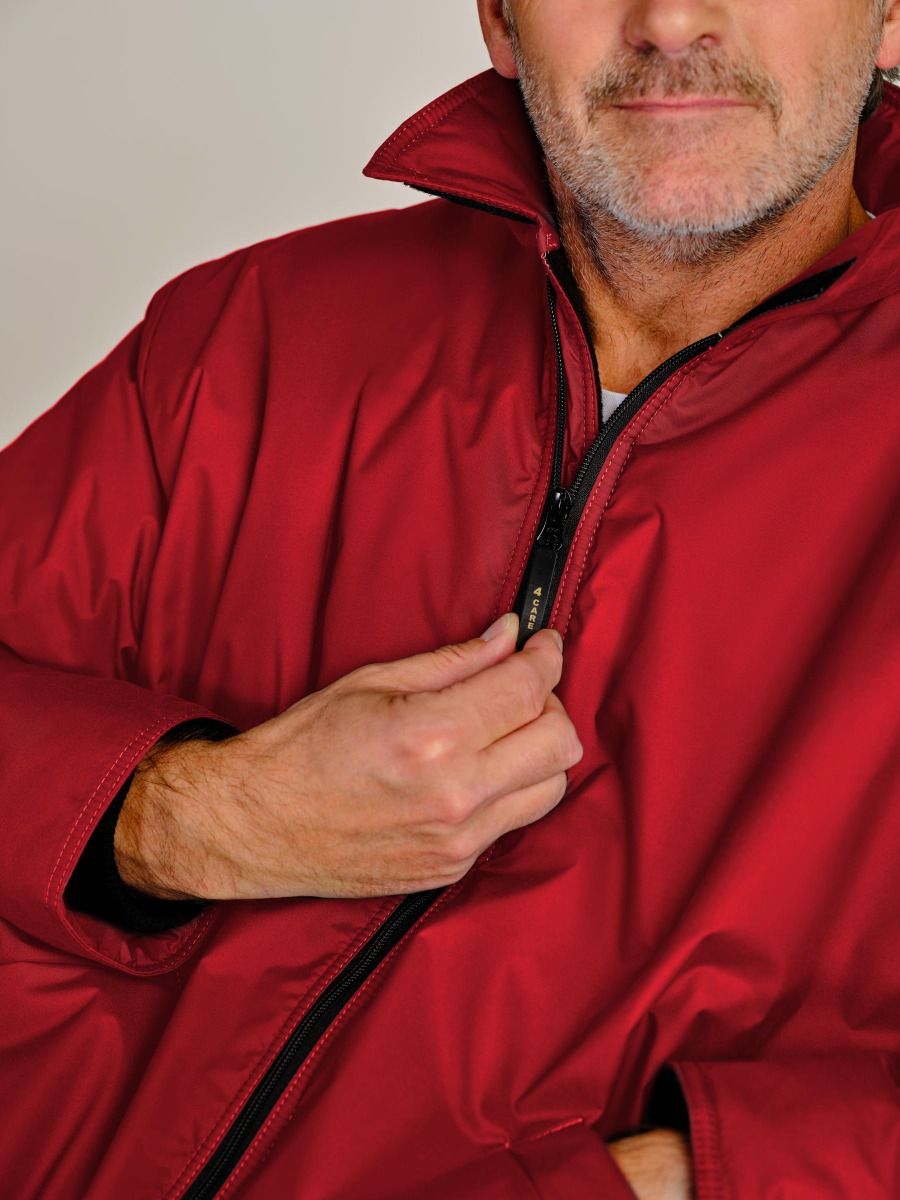 A close-up of a man wearing a red zip-up jacket, pulling the zipper with his hand. The jacket has a high collar and a smooth, slightly shiny texture.