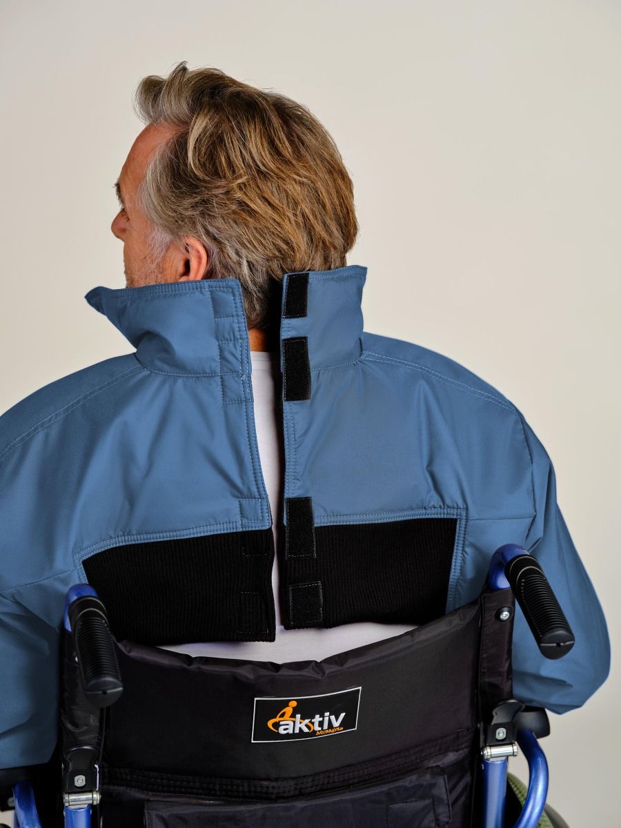 A man viewed from behind wearing a blue jacket with a black back support harness attached, seated in a wheelchair. The jacket has a high collar and black hook-and-loop fasteners on the back.