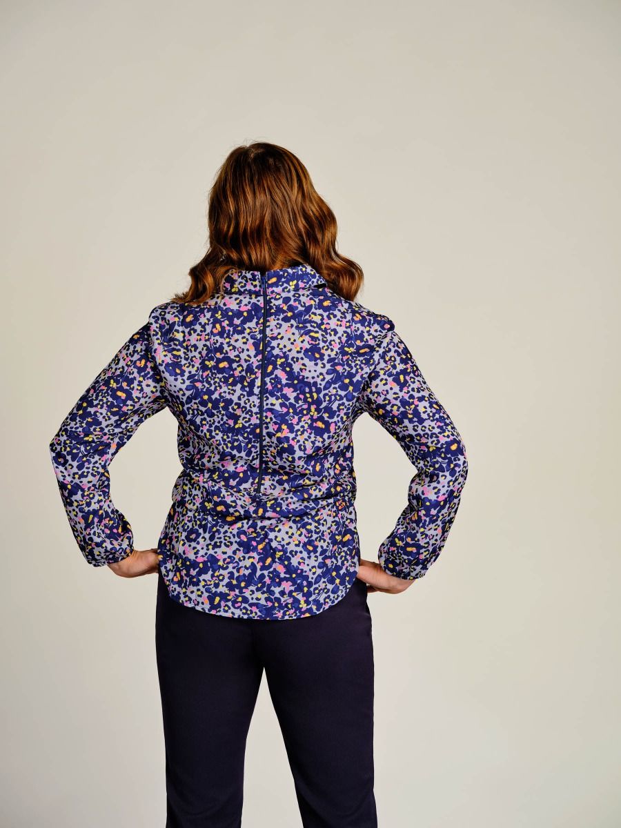 A woman viewed from behind wearing a long-sleeve button-front jacket with a dense blue, purple, and white floral pattern, paired with dark navy pants, against a plain light beige background.