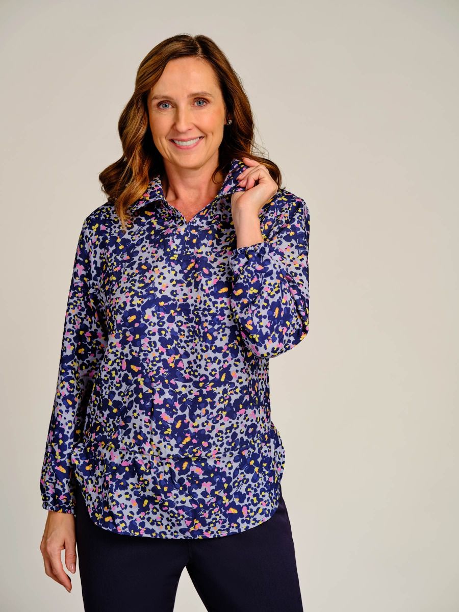 A woman modeling a long-sleeve button-up blouse with a dense abstract floral print in navy blue, purple, pink, and white, paired with dark navy pants, against a solid light gray studio background.