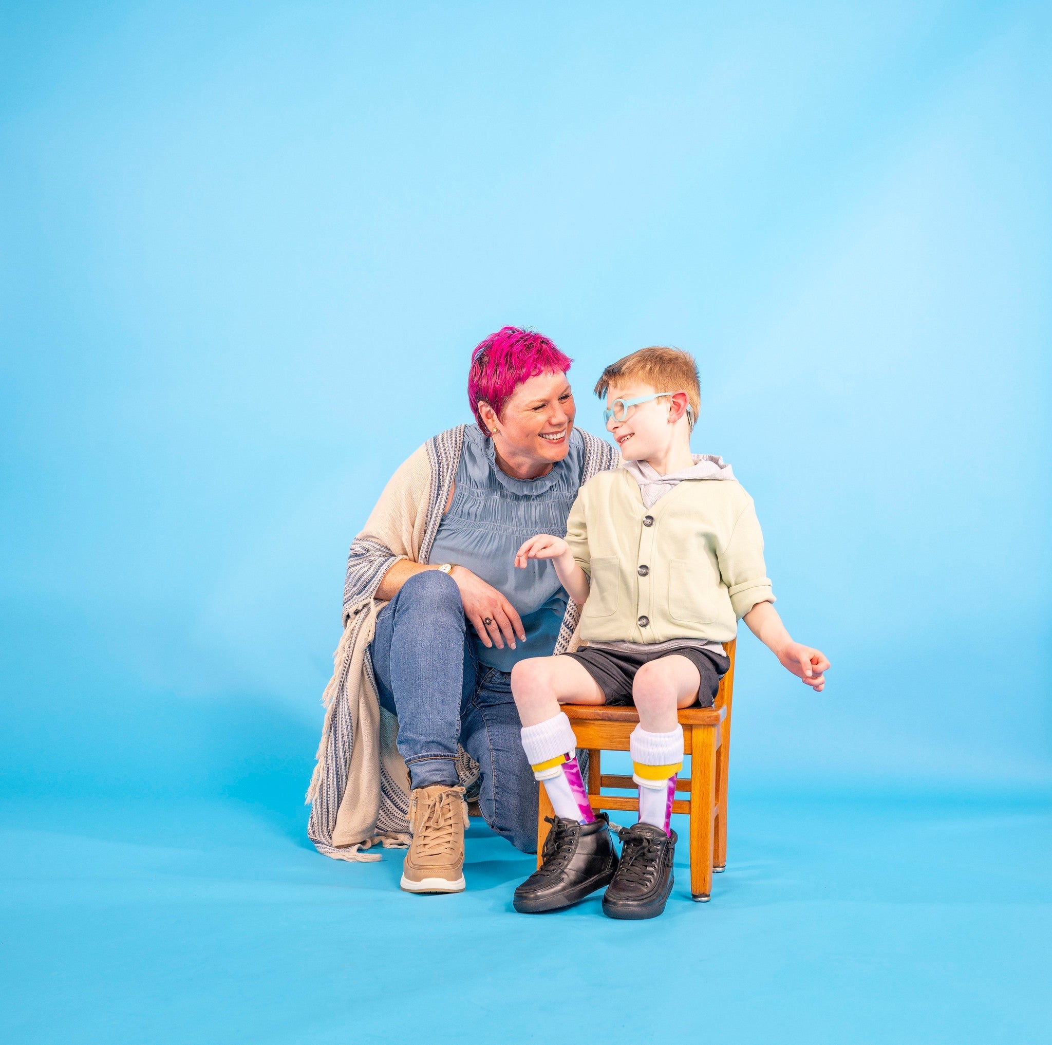A woman with bright pink hair and a young boy wearing glasses sit together on a small wooden chair against a solid blue studio background. The boy is wearing a light beige cardigan, dark shorts, white socks with colorful stripes, and black shoes.