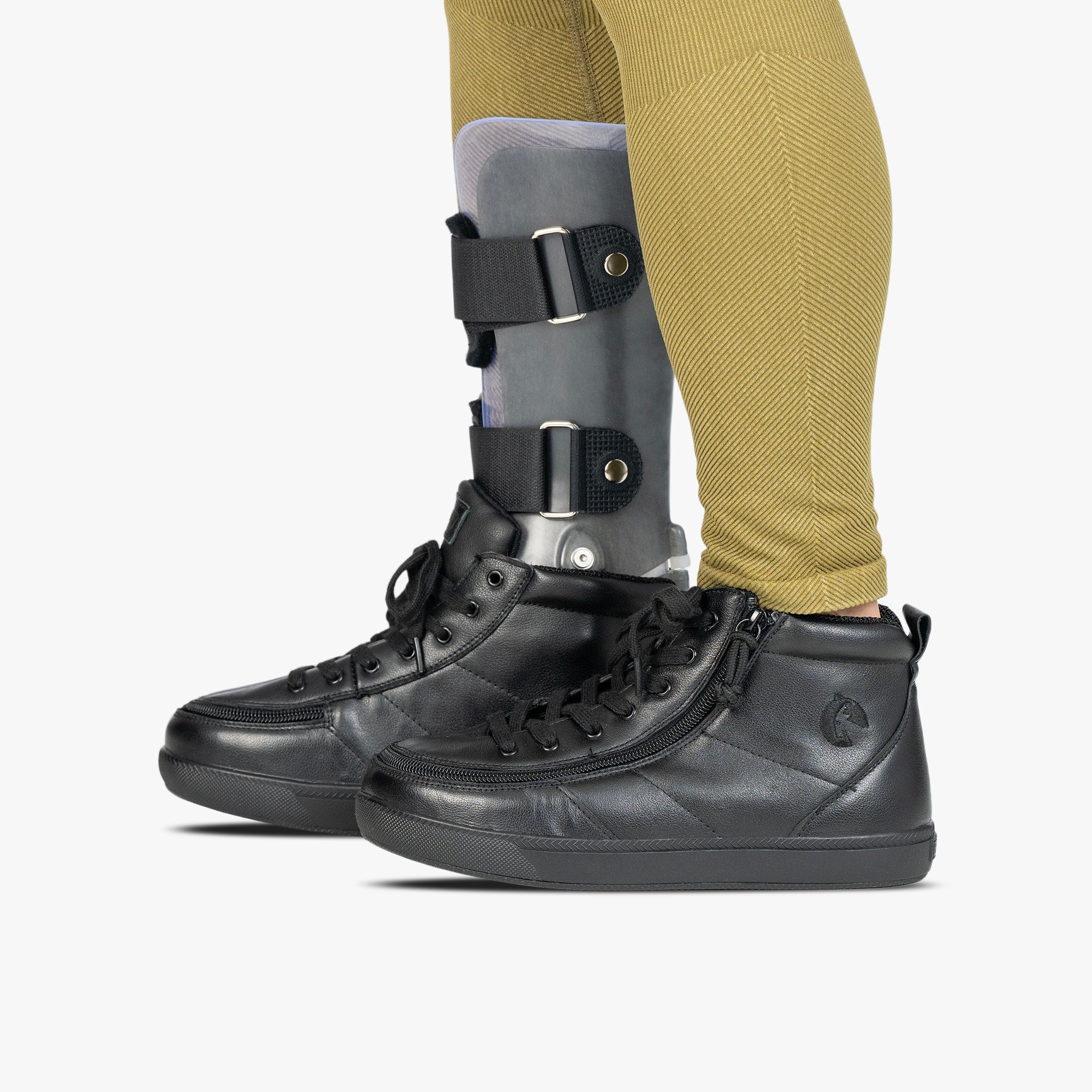 A person wearing black high-top lace-up shoes and olive yellow leggings, with a gray and black lower leg support brace visible on one leg, against a light gray background.
