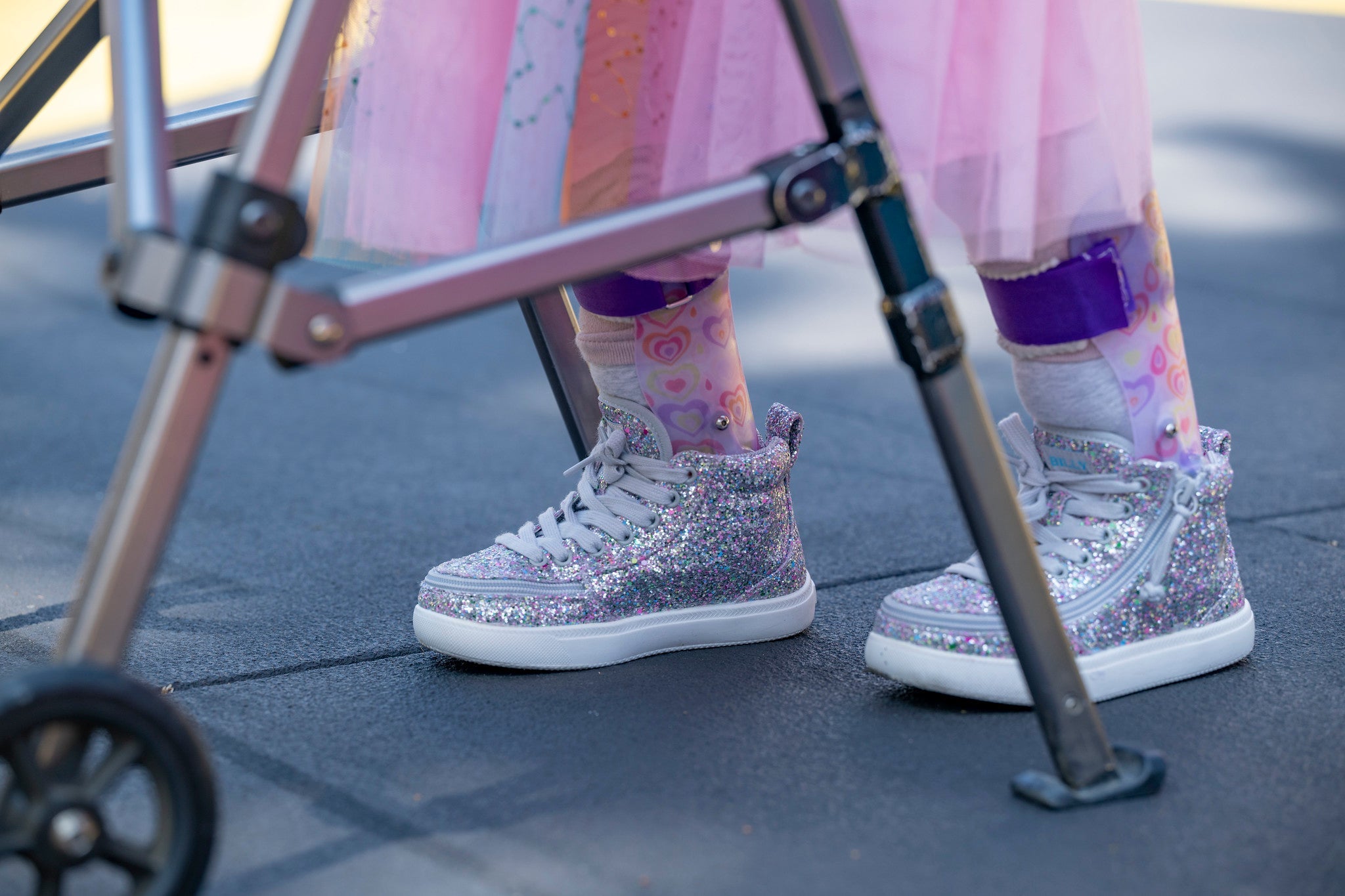 A close-up of a child's feet wearing sparkly silver high-top sneakers with white soles, pink patterned socks, and a pink tutu, standing near a metal walker outdoors.