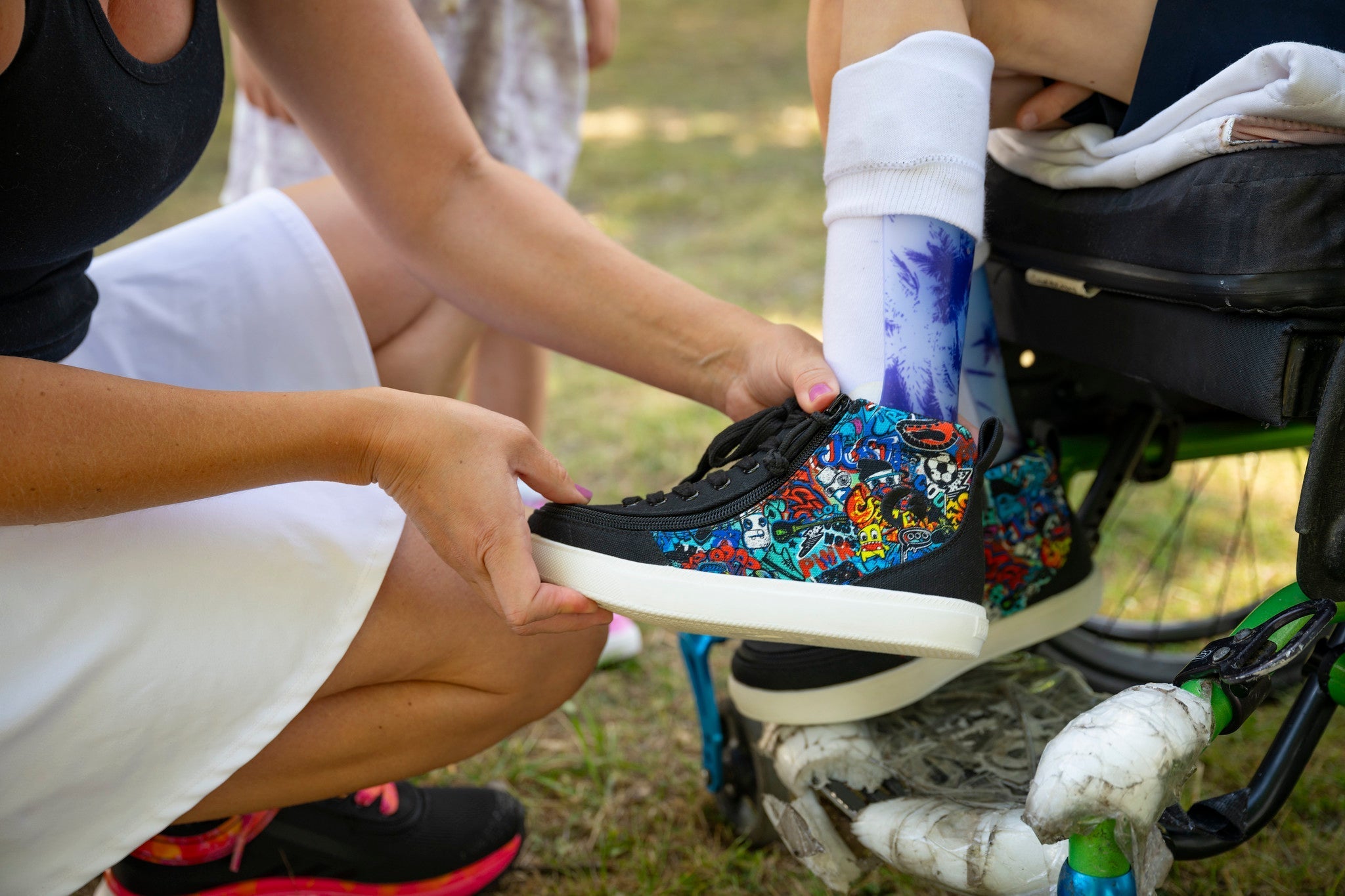 A close-up lifestyle shot of a person holding a black low-top sneaker with a vibrant multicolor sugar skull graphic print. The person is wearing a white skirt and a white sock with a blue graphic, in an outdoor setting.