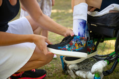 A close-up lifestyle shot of a person holding a black low-top sneaker with a vibrant multicolor sugar skull graphic print. The person is wearing a white skirt and a white sock with a blue graphic, in an outdoor setting.