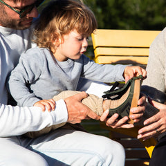 A young child sits on an adult's lap outdoors, wearing a dark green shoe while the adult helps put on the shoe. A yellow bench and blurred greenery are in the background.