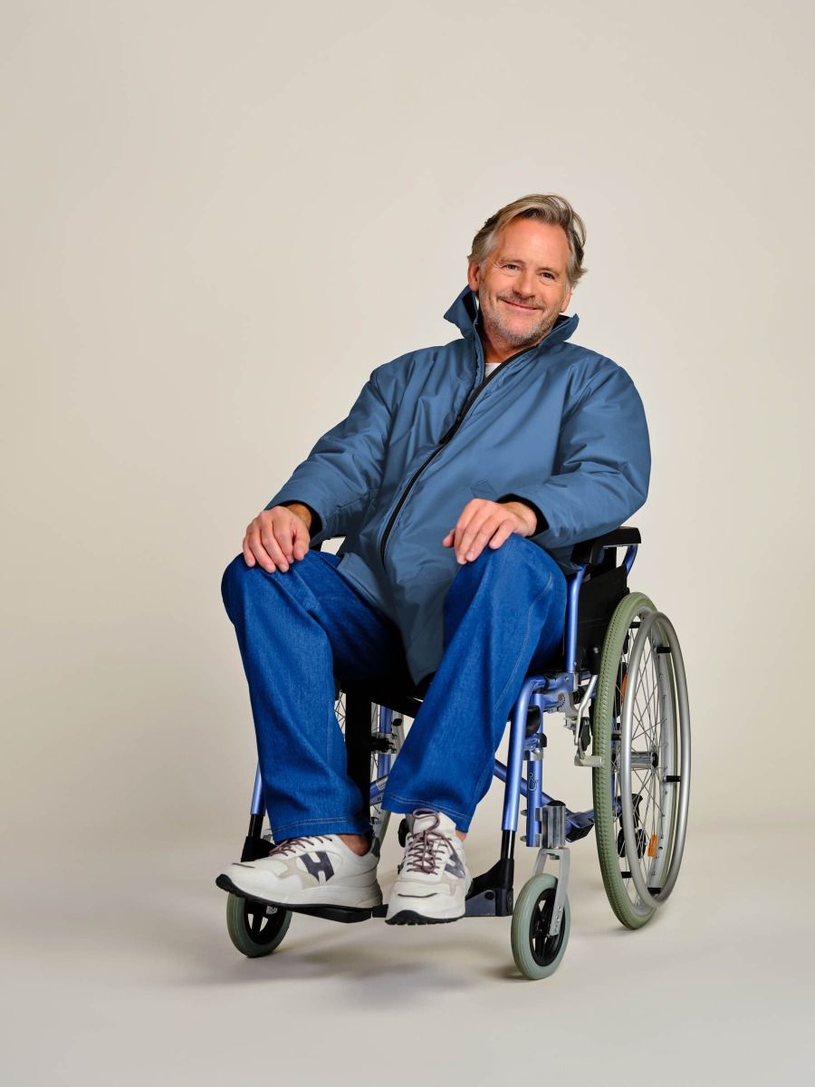 A smiling man with gray hair sits in a manual wheelchair wearing a blue zippered jacket, blue pants, and white sneakers against a plain light beige studio background.