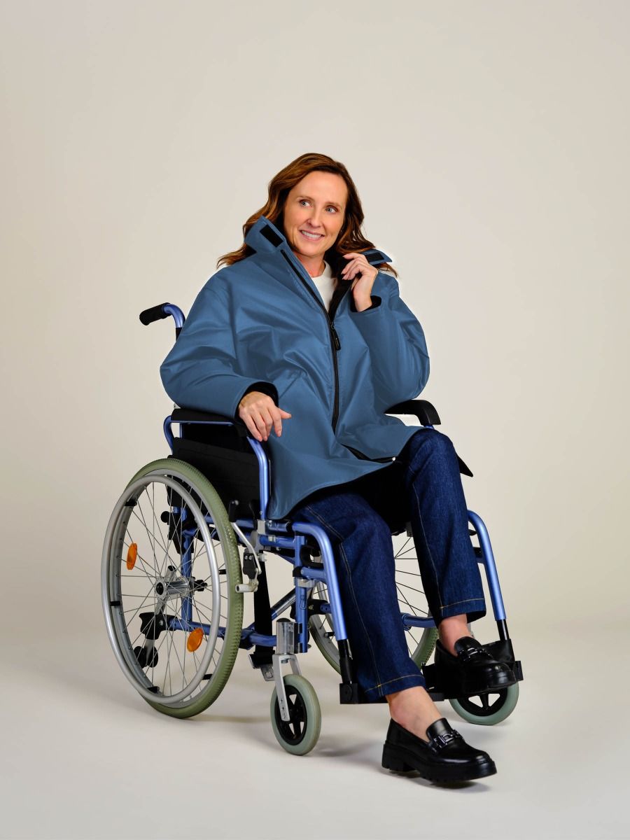A woman with long light brown hair sits in a blue manual wheelchair, wearing a blue zippered jacket, dark blue jeans, and black loafers against a plain light gray studio background.