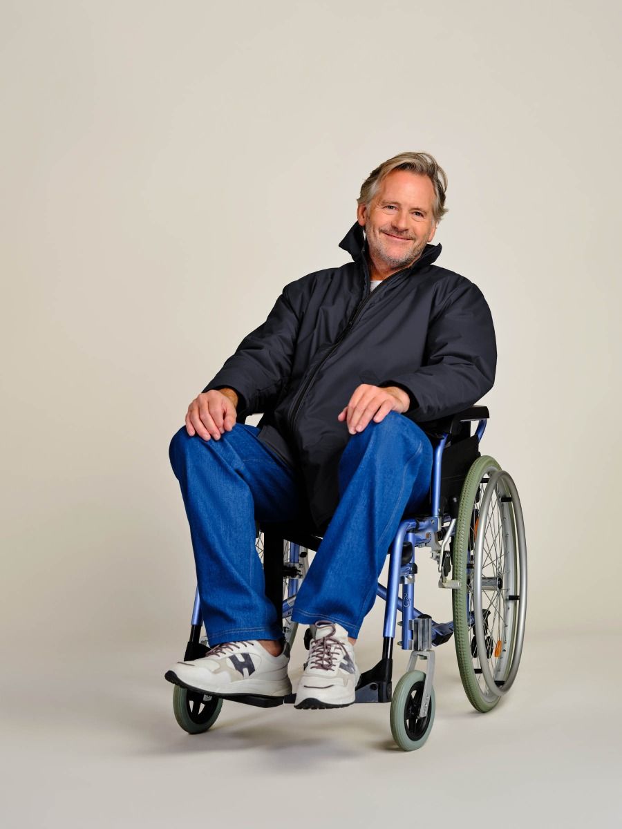 A smiling man with blond hair sits in a blue manual wheelchair, wearing a black zip-up jacket, blue jeans, and white sneakers, photographed against a plain light beige studio background.