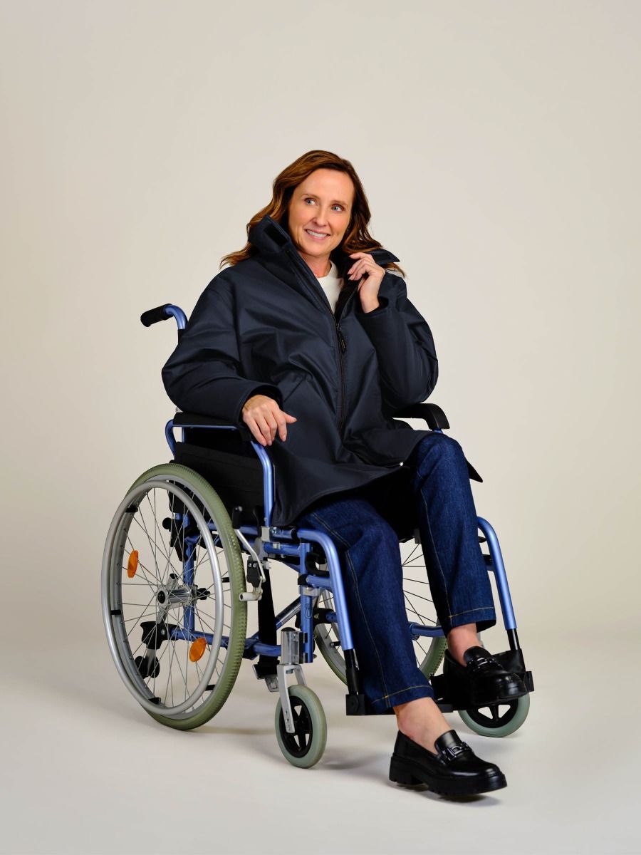 A woman with shoulder-length brown hair sits in a blue manual wheelchair, wearing a dark navy blue puffer jacket, blue jeans, and black leather loafers against a plain light beige studio background.