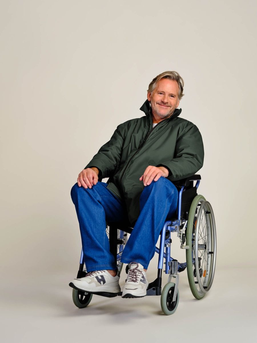 A smiling man with gray hair sits in a blue manual wheelchair, wearing a dark green zip-up jacket, blue jeans, and white New Balance sneakers against a plain light gray studio background.
