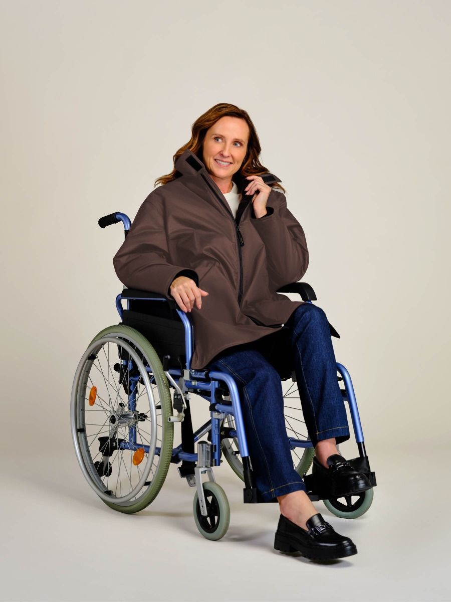 A woman with shoulder-length brown hair sits in a blue manual wheelchair, wearing a brown hooded jacket, blue jeans, and black loafers against a plain light beige studio background.