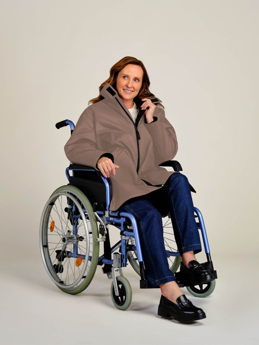 A woman sitting in a blue manual wheelchair wearing a brown zippered jacket, blue jeans, and black loafers, photographed against a plain light beige studio background.