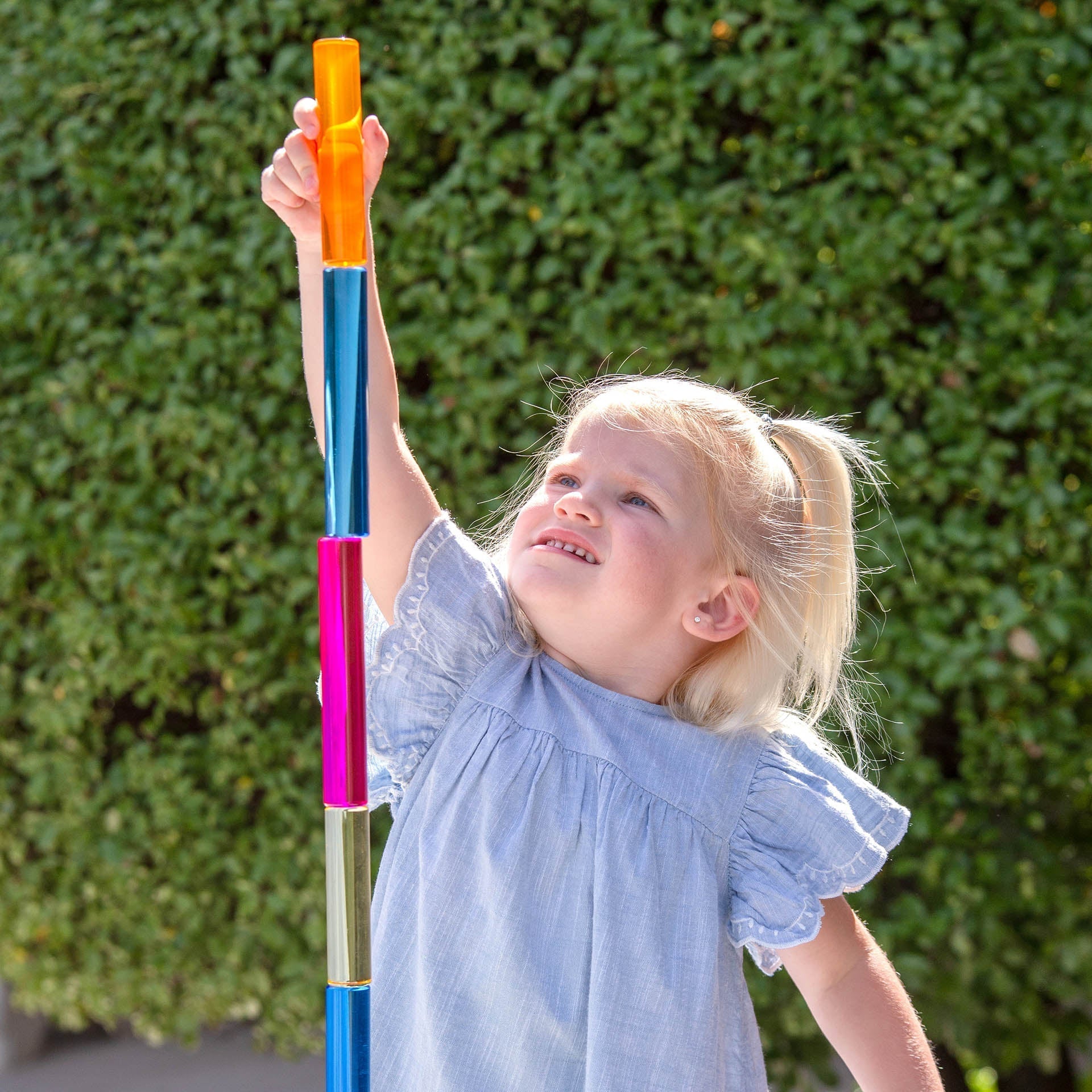 Child holding a colorful stick against a green hedge background