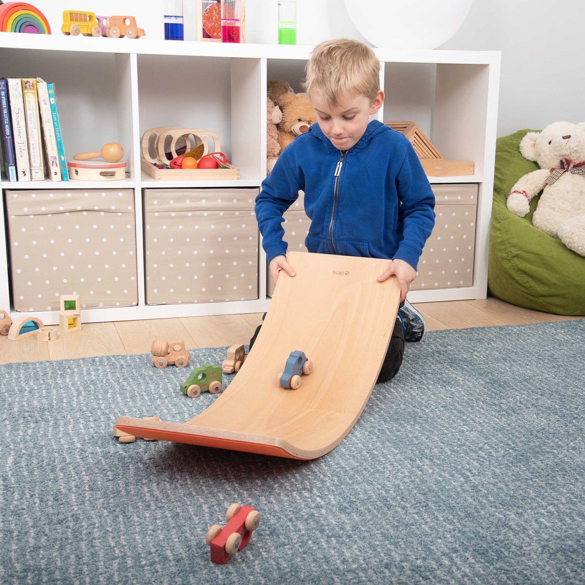 Child playing with a wooden toy ramp in a room with shelves and toys.