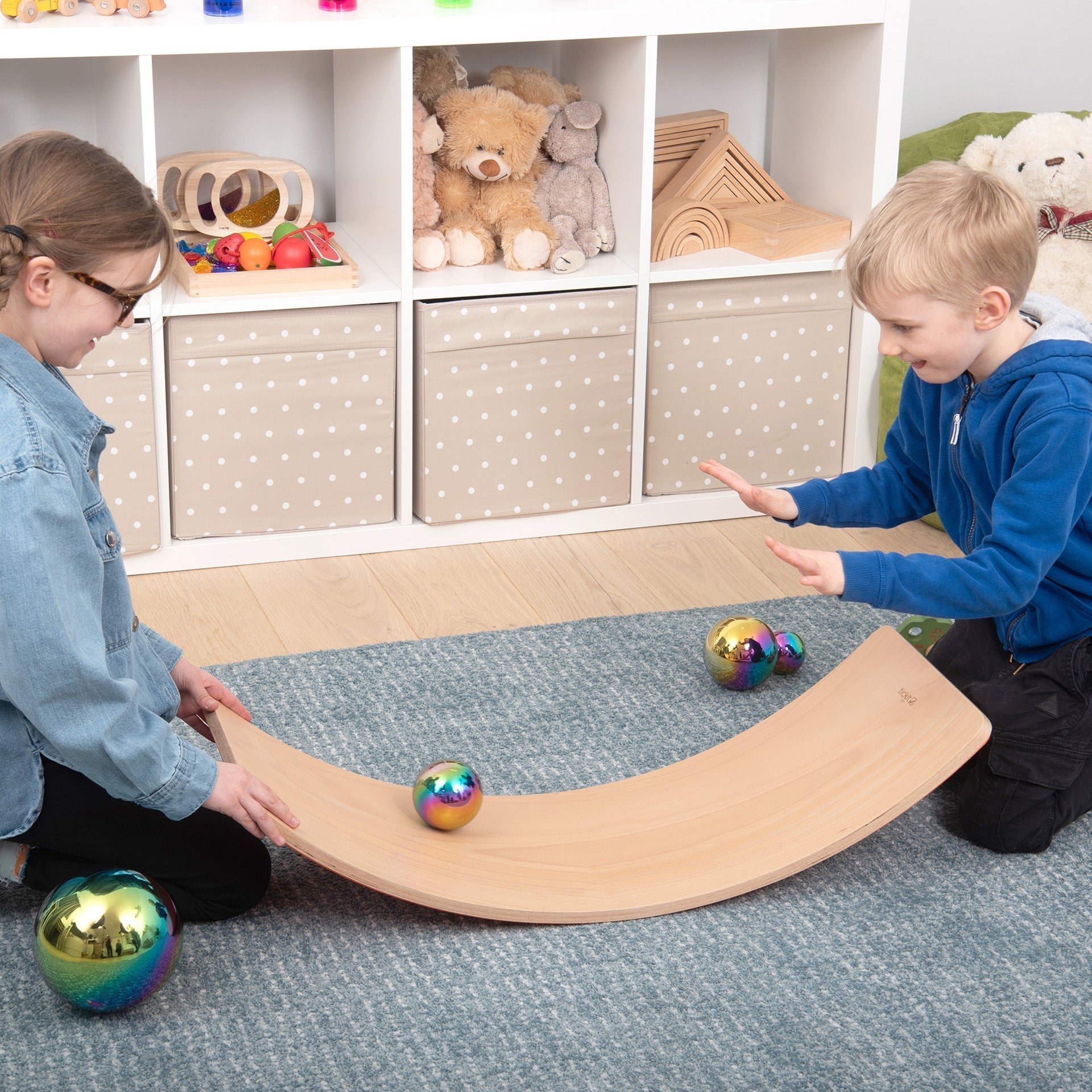 Two children playing with colorful balls on a wooden seesaw in a room with shelves and toys.