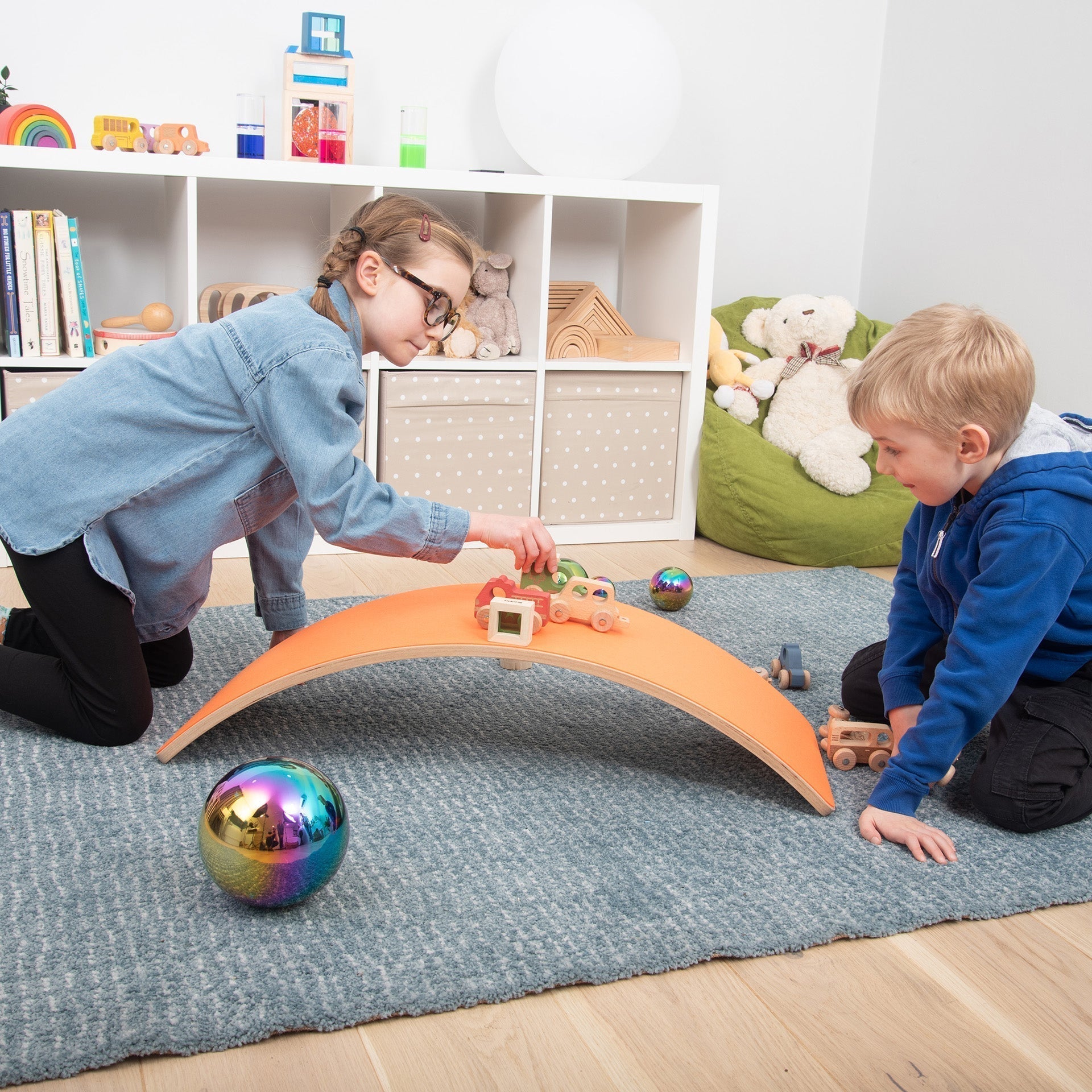 Two children playing with a toy track in a room with shelves and toys.