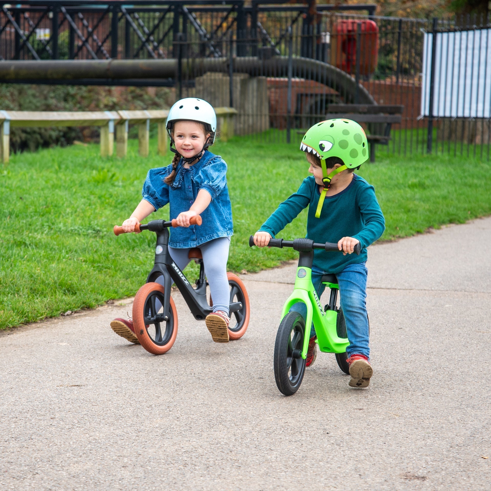 Two children riding balance bikes on a path with grass and playground equipment in the background.