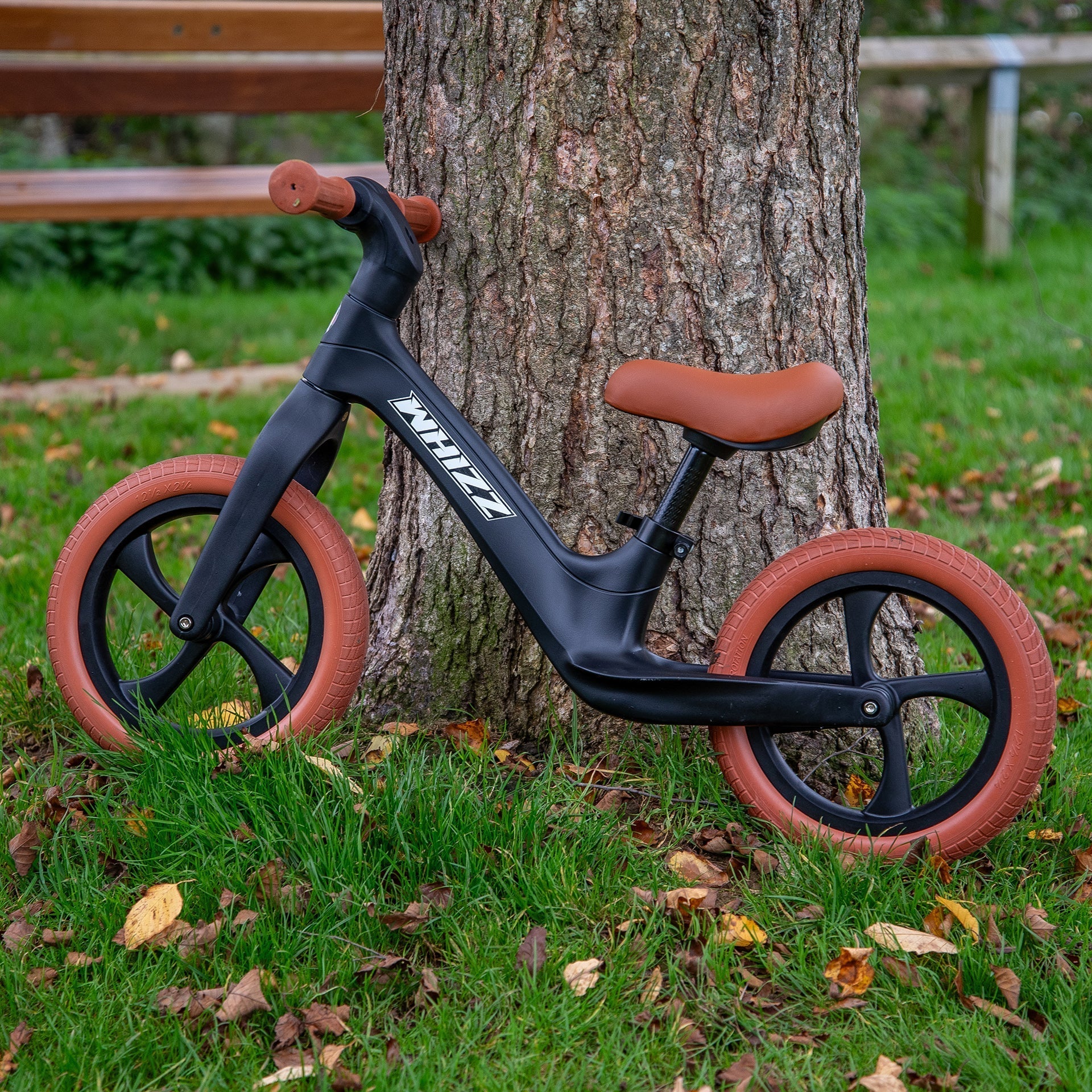 Children's balance bike with brown wheels leaning against a tree in a park setting.
