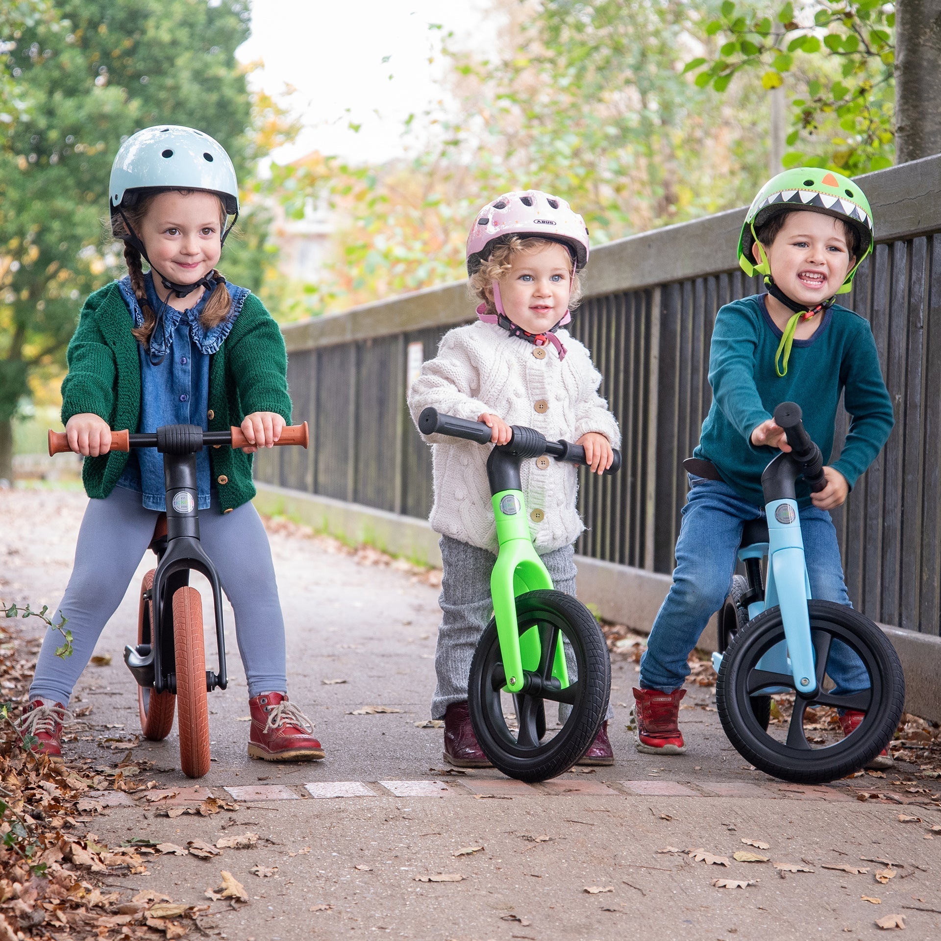 Three children riding balance bikes on a path with trees in the background