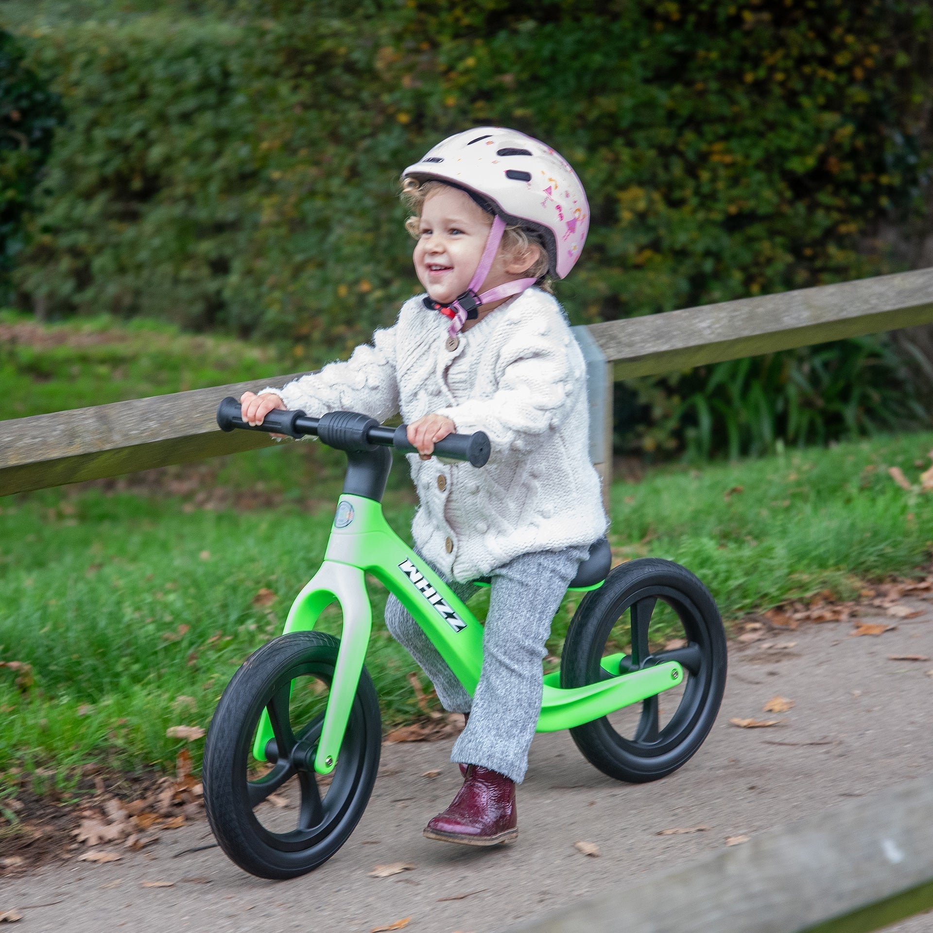 Child riding a green balance bike outdoors on a path with grass and wooden fence in the background.