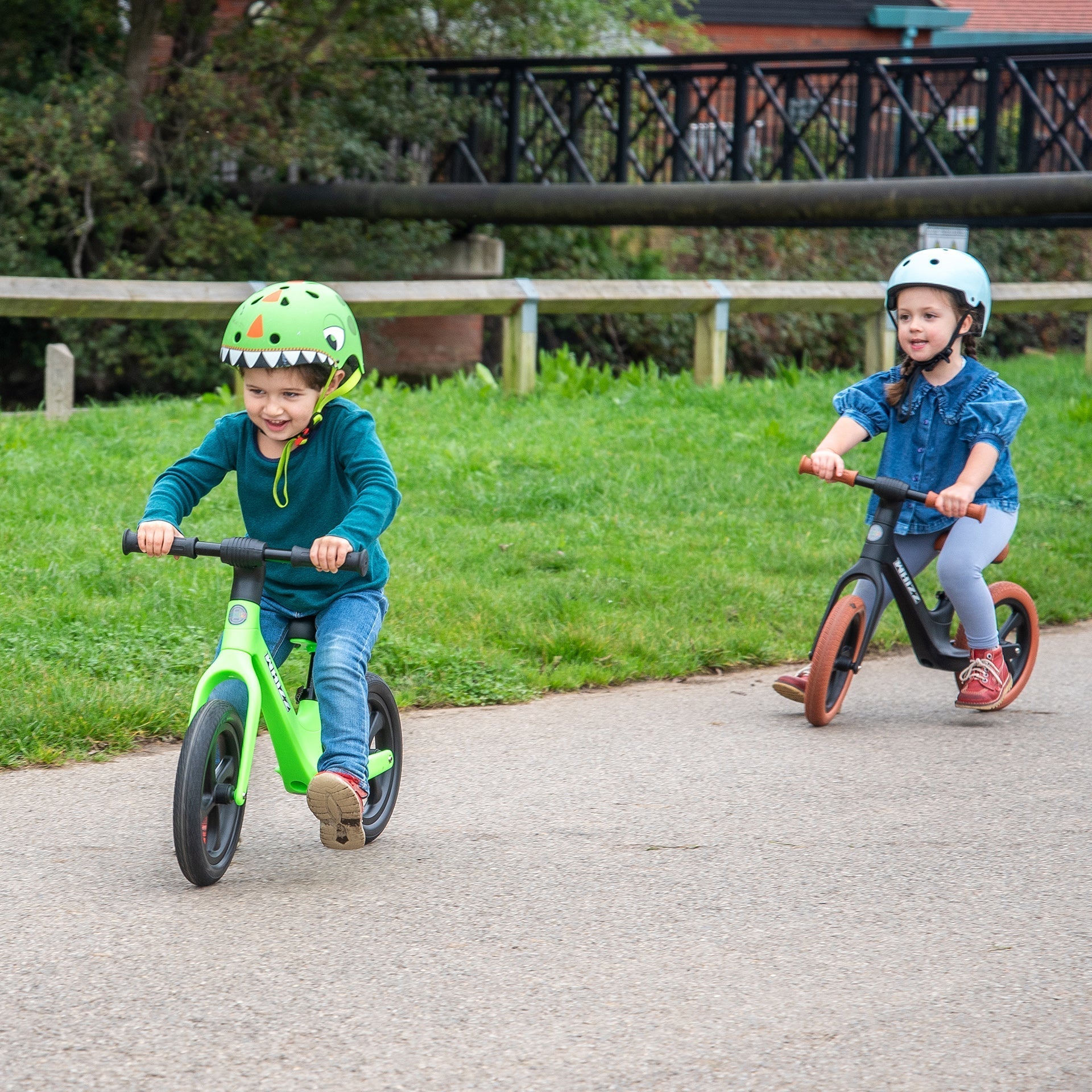 Two children riding balance bikes on a path with greenery in the background