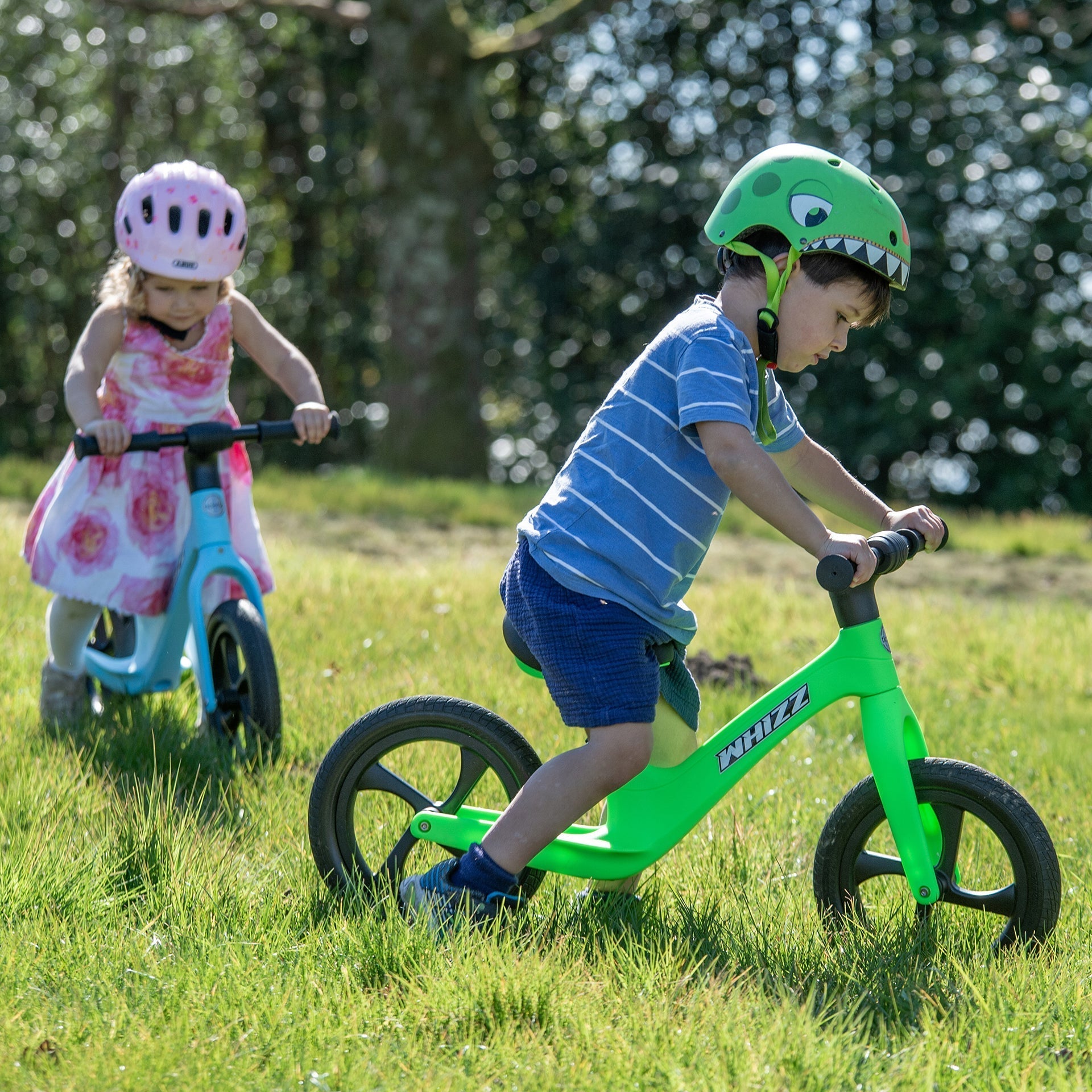 Two children riding balance bikes in a grassy area with trees in the background