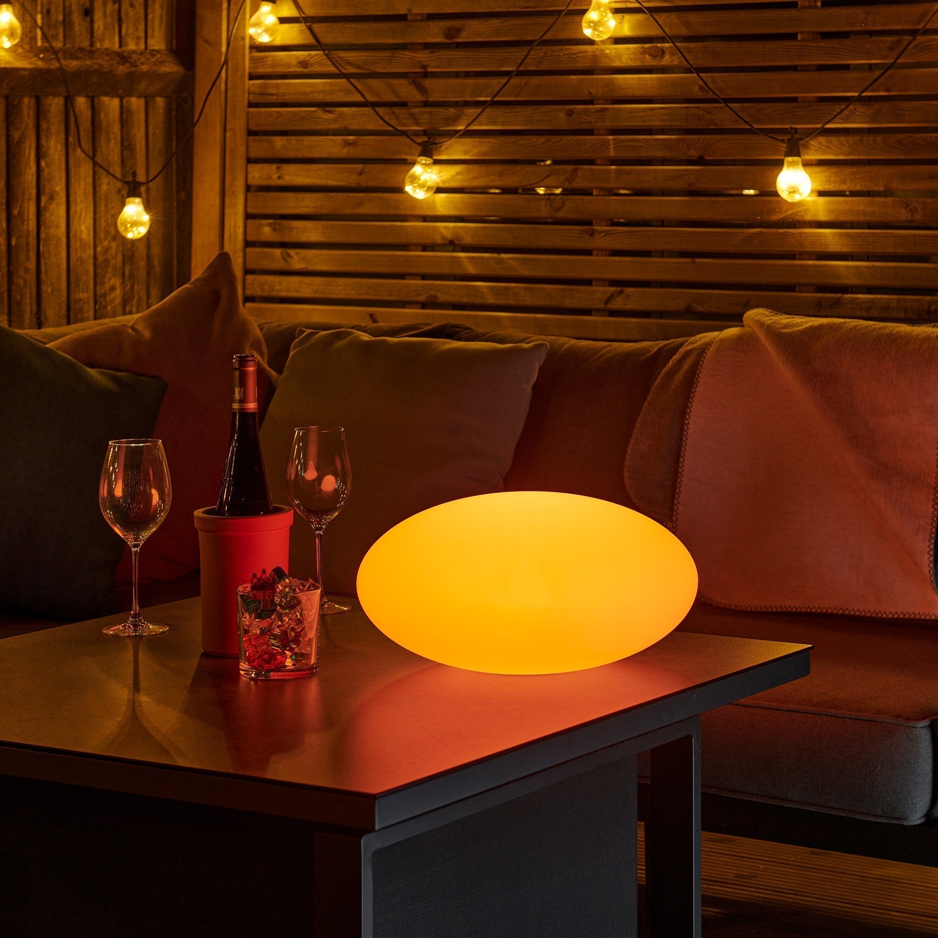 Cozy indoor setting with a lit table lamp, wine glasses, and a bottle on a wooden table.