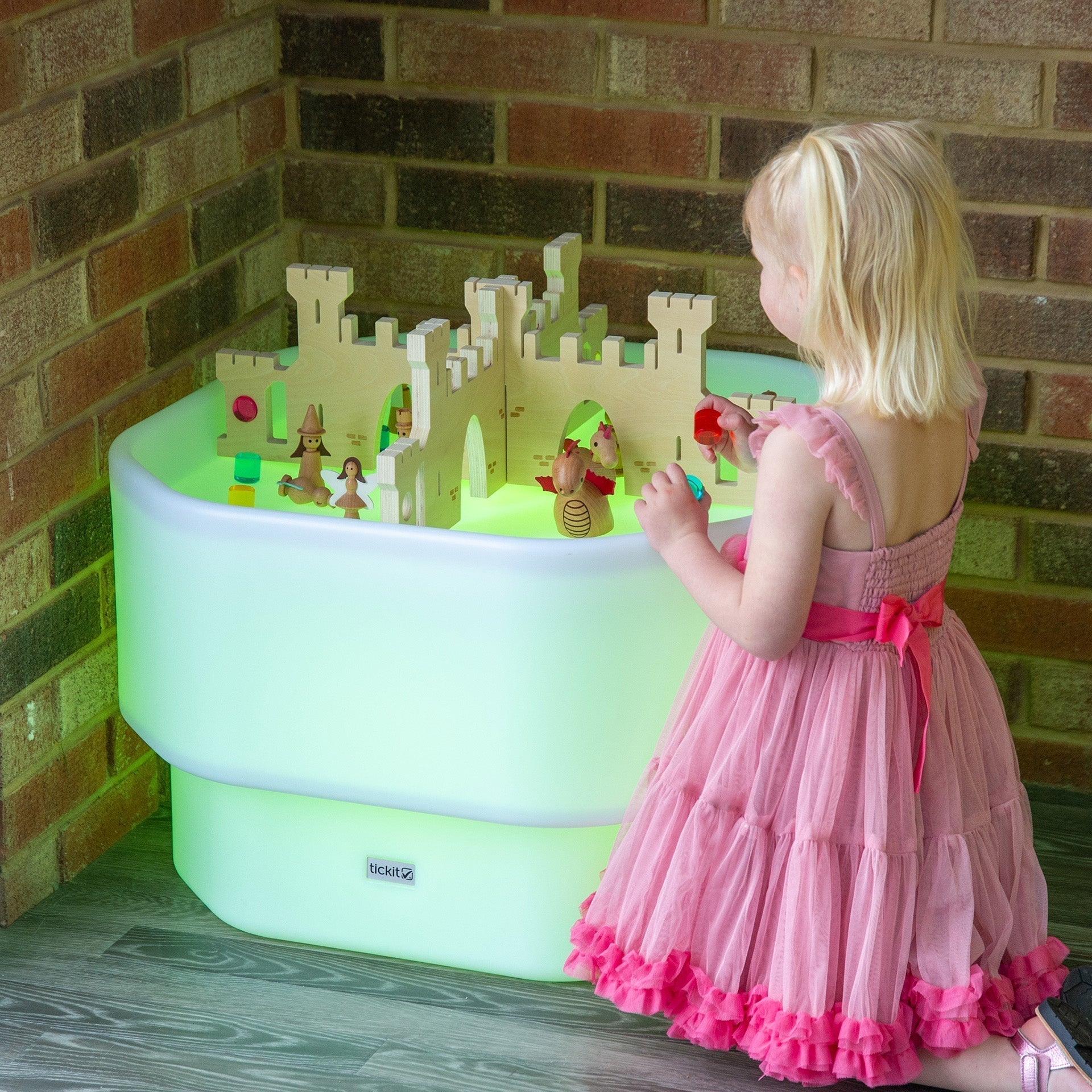 Child in a pink dress playing with a toy castle in a green playpen against a brick wall.