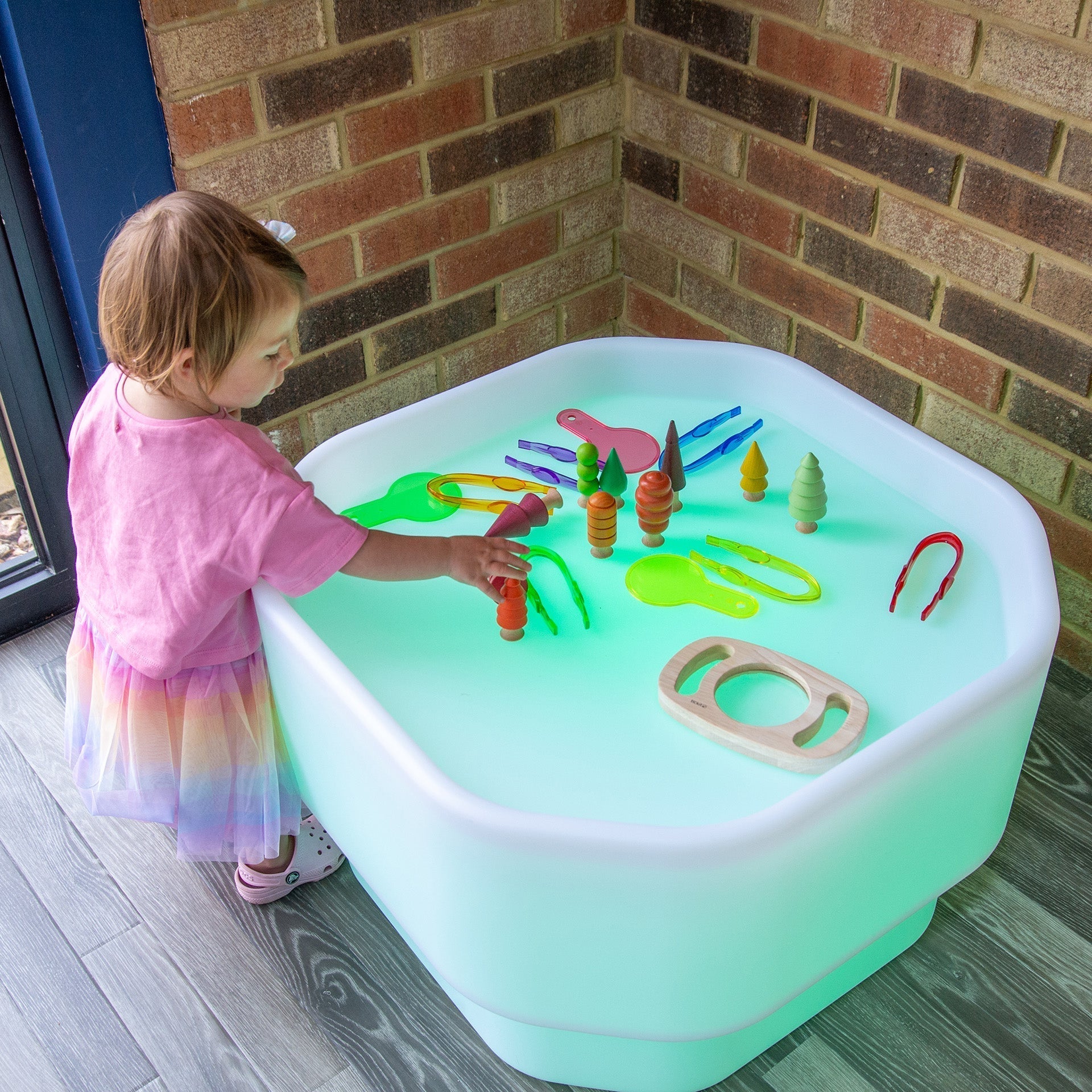 Child playing with toys on a colorful LED play table against a brick wall.