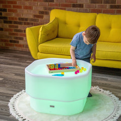 Child playing with toys on a glowing green table in a room with a yellow couch and brick wall.