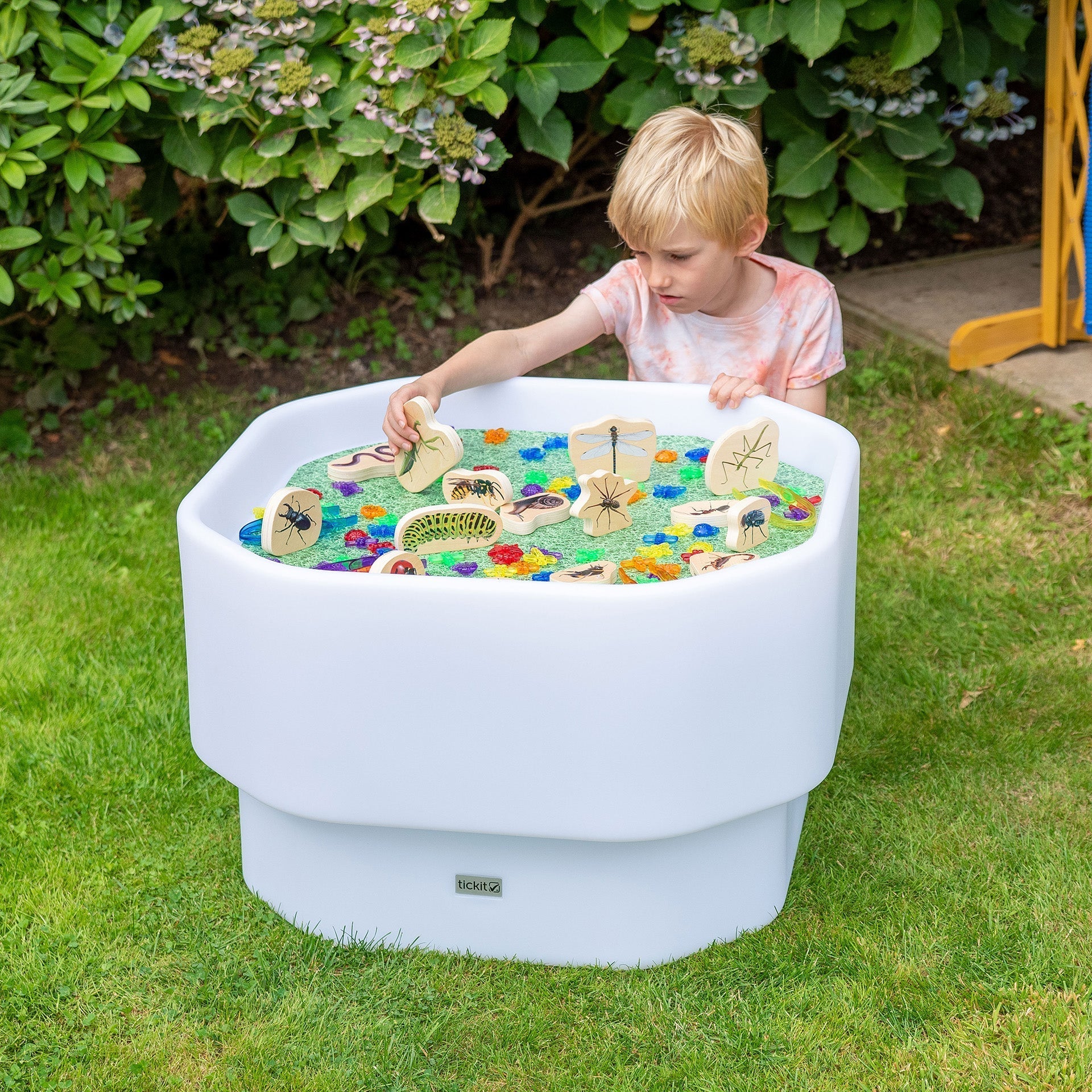 Child playing with a toy boat in a white outdoor play table on a grassy area.