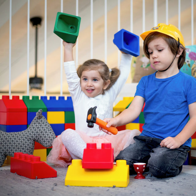 Two children playing with colorful toy building blocks and a small toy hammer in a bright indoor play area. The child on the left holds a green block above her head, and the child on the right wears a yellow hard hat while holding an orange-handled hammer.