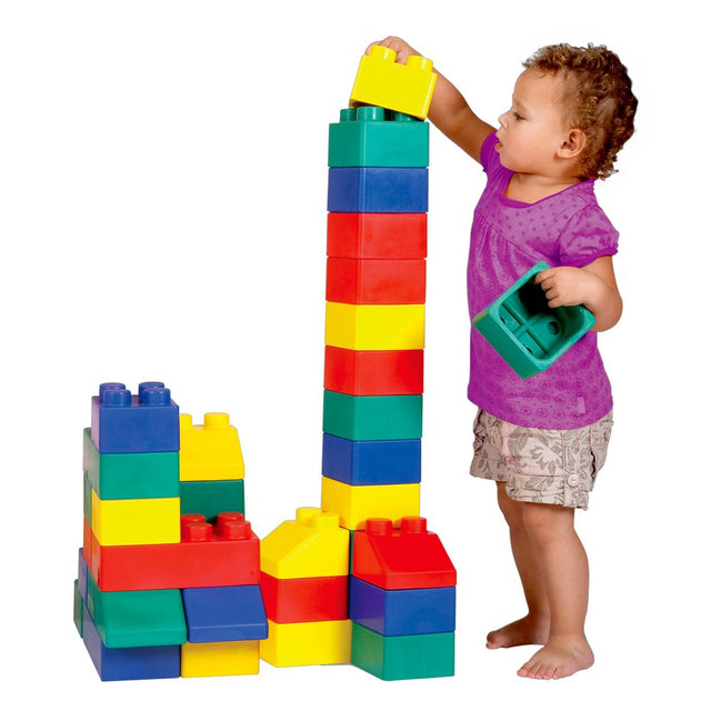 A toddler stands next to a large stack of colorful oversized building blocks on a white background, placing a yellow block on top of a tall tower while holding a teal block in one hand.