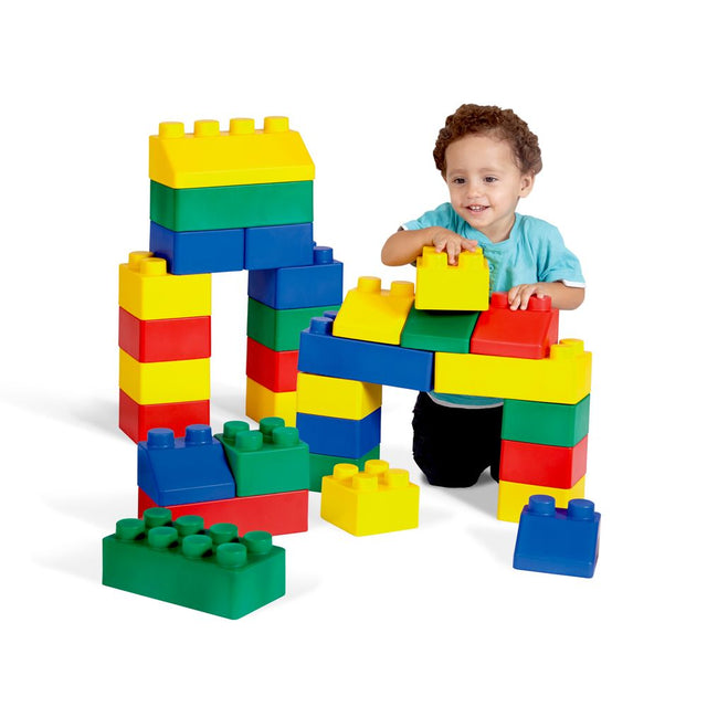 A young child with curly brown hair wearing a light blue t-shirt and black pants is playing with a set of colorful oversized building blocks, including yellow, green, blue, and red pieces, on a clean white background.