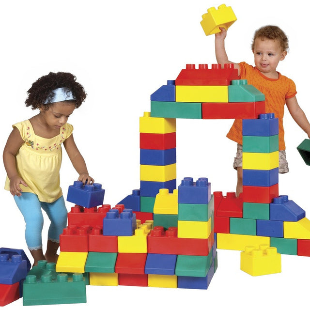 Two young children playing with large colorful foam building blocks on a white background. One child in a yellow top and blue pants is placing a blue block, while the other in an orange shirt holds up a yellow block above a structure made of red, yellow, blue, and green blocks.