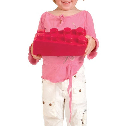 A young child wearing a pink long-sleeve top and white pants holds a large, oversized bright pink toy brick with studs against a plain white background.